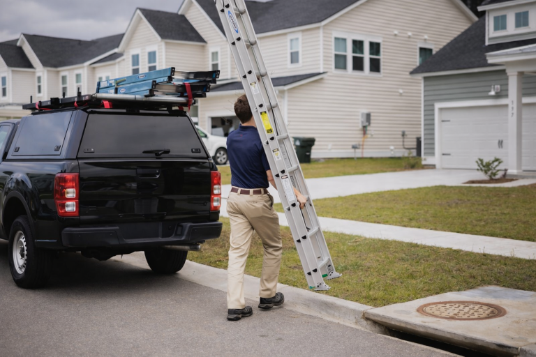 A wildlife removal technician carries a ladder towards a house.